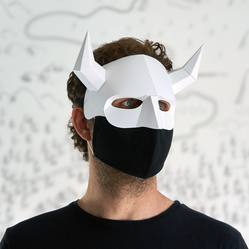 A man wearing a white paper demon or oni-style horned mask and a black fabric face mask, standing against a light wall with faint hand-drawn line art.