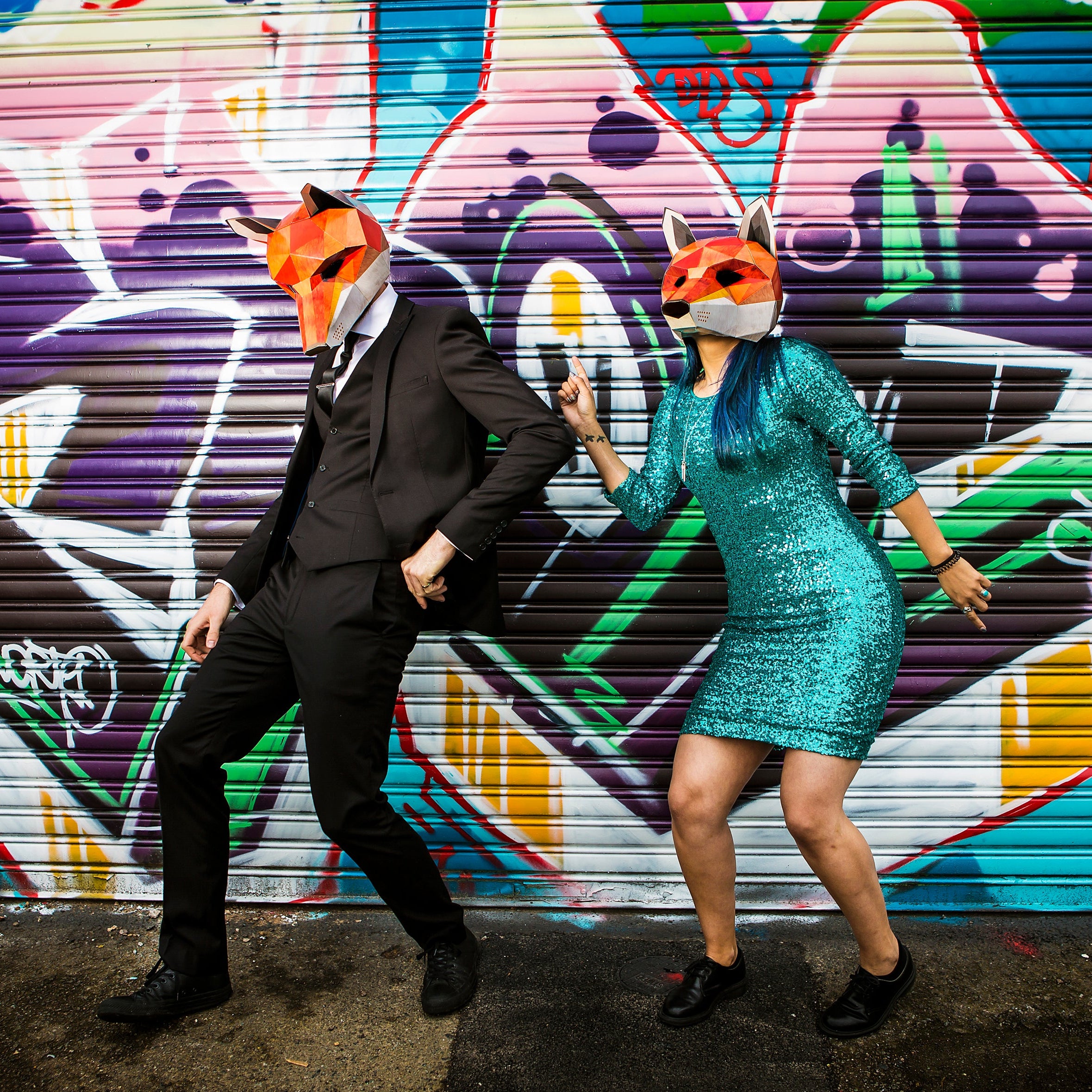 Two people wearing fox masks dancing in front of a colorful graffiti wall.