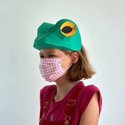 Child wearing a green frog hat with a yellow eye patch and a pink checkered mask on a white background