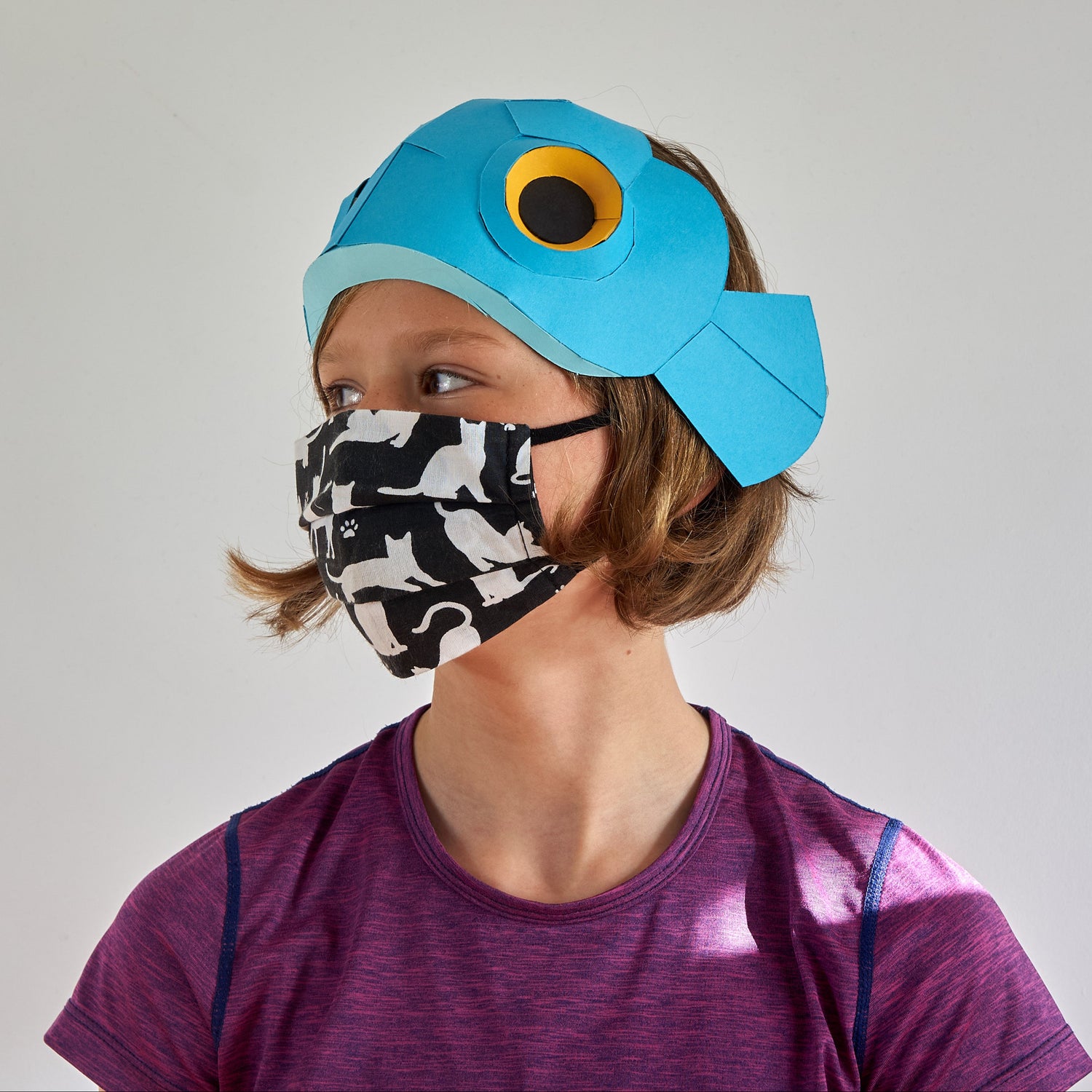 Person wearing a blue fish hat and a patterned face mask on a plain background