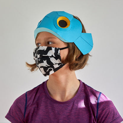 Person wearing a blue fish hat and a patterned face mask on a plain background