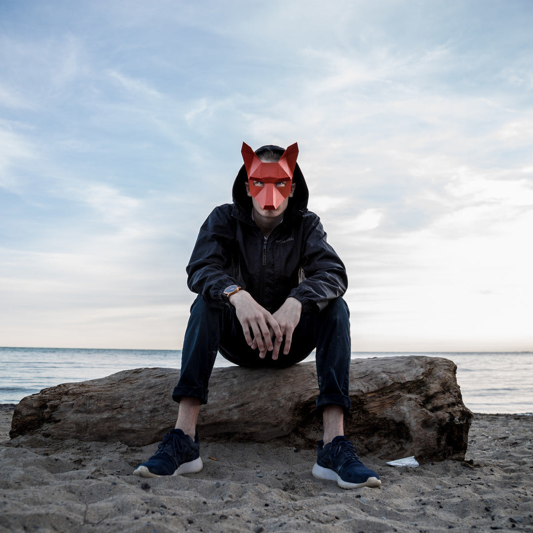 A person wearing a wintercroft fox mask sitting on a log by the beach